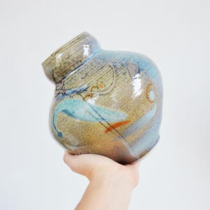 May include: A rounded ceramic vase with a glossy finish, featuring a mottled blend of tan, brown, and blue tones. The surface is decorated with abstract painted shapes in light blue, orange, and dark brown, including thin lines and circular motifs.