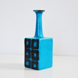 K&ouml;nnte beinhalten: Eine kleine, blaue Keramikvase mit quadratischem Sockel und einem hohen, d&uuml;nnen Hals. Die Vase ist mit schwarzen geometrischen Formen verziert.