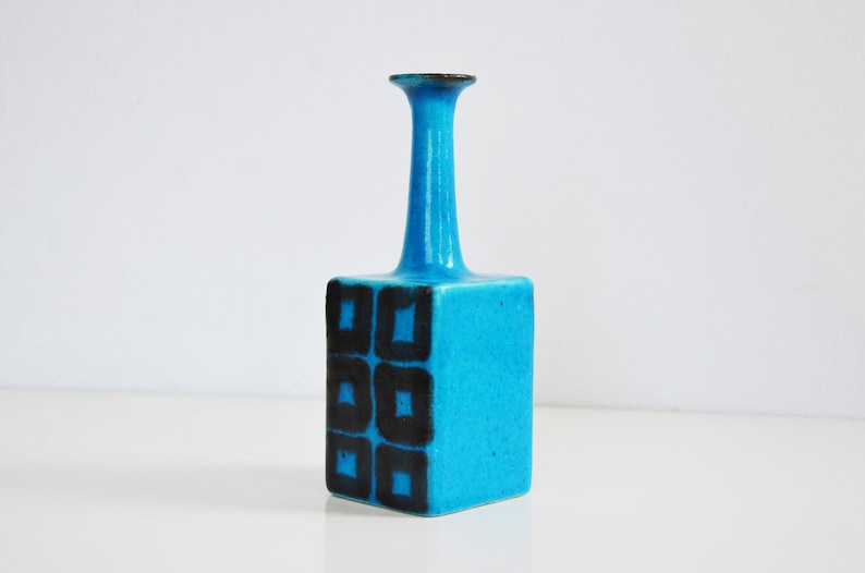 K&ouml;nnte beinhalten: Eine kleine, blaue Keramikvase mit quadratischem Sockel und einem hohen, d&uuml;nnen Hals. Die Vase ist mit schwarzen geometrischen Formen verziert.