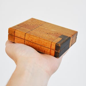 May include: A rectangular wooden box with a hinged lid, held in a hand. The box is made of light brown wood with darker brown accents and decorative lines. The box is approximately 4 inches long.