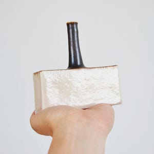 Puede incluir: Un tapón de botella de cerámica con base rectangular y cuello curvo marrón oscuro. La base es blanquecina con superficie texturizada y borde marrón. El tapón se sostiene en una mano sobre un fondo blanco.