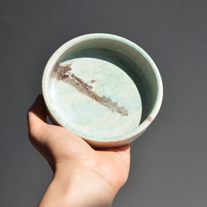 May include: A small, round ceramic bowl with a light teal interior and a white exterior. A brown, linear design is painted on the inside. The bowl is held in a hand against a grey background.