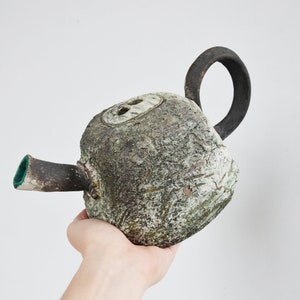 May include: A rustic, hand-crafted ceramic teapot with a textured, green and white glaze. The teapot has a unique, rounded shape and a black handle.