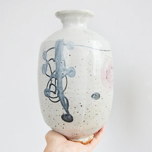 Könnte beinhalten: Eine weiße Keramikvase mit gesprenkelter Glasur und abstrakten, in Blau und Schwarz gemalten Designs. Die Vase hat einen runden Körper und einen schmalen Hals.