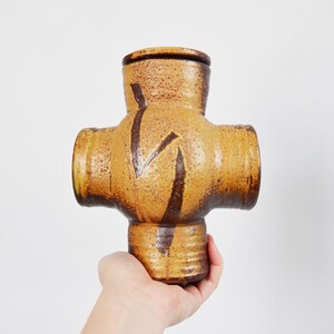 May include: A handmade, cross-shaped ceramic vase in shades of brown and tan. The vase has four cylindrical openings and is decorated with dark brown zigzag patterns. The vase is held by a hand against a white background.