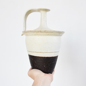 May include: A handmade ceramic pitcher with a handle and a lid. The upper portion is off-white, while the lower section is dark brown. The pitcher is held by a hand against a white background. The height is approximately 30 cm.