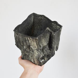 Puede incluir: Un recipiente de forma cuadrada con un exterior oscuro y texturizado que imita la apariencia de roca rugosa y desgastada. La superficie presenta grietas profundas y crestas irregulares en tonos carbón, negro y gris apagado.