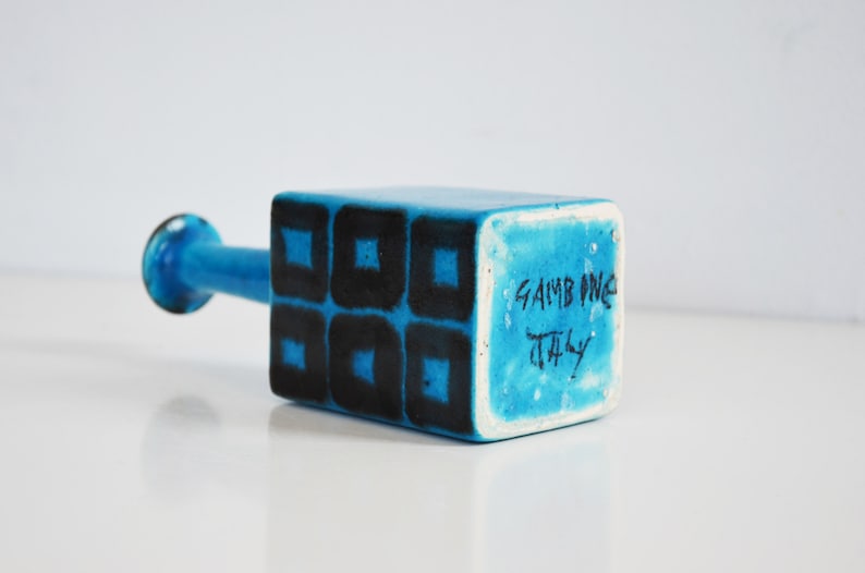 K&ouml;nnte beinhalten: Eine blaue Keramikvase mit quadratischem Sockel und zylindrischem Hals. Die Vase hat ein geometrisches Muster aus schwarzen Quadraten auf blauem Hintergrund. Der Sockel der Vase ist mit "Gambone Italy" gekennzeichnet.
