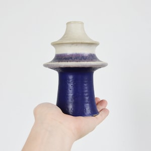 May include: A white and purple ceramic vase with a unique, tiered design. The vase has a wide, flat base and a narrow, cylindrical neck.