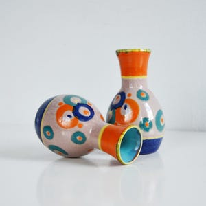 Op de afbeelding: Twee kleine, decoratieve keramische vazen. Eén vaas ligt op zijn kant en de andere staat rechtop. Beide vazen hebben een lichtroze basiskleur met blauwe, oranje en turquoise cirkelvormige ontwerpen. De randen en halzen zijn oranje.