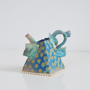 Puede incluir: Una tetera de cerámica única con un estampado de lunares azules y amarillos. La tetera tiene forma triangular con asa y boquilla curvadas. Los bordes están adornados con un borde blanco texturizado. La tetera es una pieza de arte decorativa.