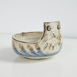 May include: A small, white ceramic bowl with a blue and brown glaze. The bowl has a unique design with an owl face on the front.