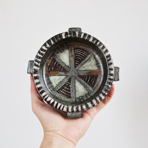 May include: A round ceramic bowl with four small handles on the rim. The interior features a geometric pattern of alternating dark brown and light green triangular segments with etched lines. The rim has a dark brown and white striped design.