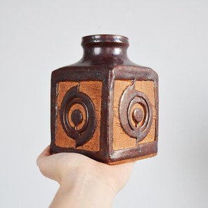 Puede incluir: Un jarrón de cerámica marrón y beige con un diseño geométrico. El jarrón tiene forma cuadrada y está decorado con círculos concéntricos.