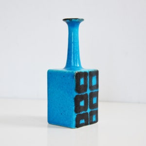 K&ouml;nnte beinhalten: Eine kleine, blaue Keramikvase mit quadratischem Sockel und einem hohen, d&uuml;nnen Hals. Die Vase hat ein geometrisches Muster aus schwarzen Quadraten auf der Vorderseite.
