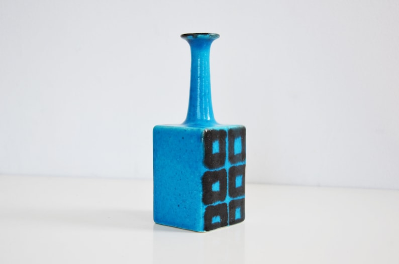 K&ouml;nnte beinhalten: Eine kleine, blaue Keramikvase mit quadratischem Sockel und einem hohen, d&uuml;nnen Hals. Die Vase hat ein geometrisches Muster aus schwarzen Quadraten auf der Vorderseite.