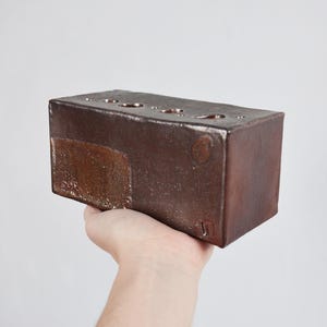 May include: A rectangular, brown ceramic vase with several circular openings on top. The vase has a textured surface and is held in a hand against a white background. The vase has a unique, handmade appearance.