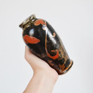 May include: A small ceramic vase with a glossy black glaze featuring hand-painted orange floral motifs and brown earthy accents. The vase has a narrow neck, a flared rim, and a tapered body with a smooth, reflective surface finish throughout.