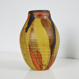 Op de afbeelding: Een handgemaakte keramische vaas met een gele basis en een abstract bruin, zwart en oranje geschilderd ontwerp. De vaas heeft een afgeronde body en een smalle hals. De vaas is ongeveer 20 cm hoog.