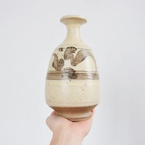 May include: A beige ceramic vase with a rounded body and a narrow neck. The vase features brown abstract designs and horizontal bands. The base is a darker shade of brown. The vase is held by a person's hand.