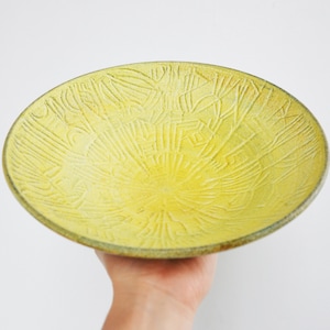 May include: A handmade, yellow ceramic bowl with a textured surface. The bowl has a circular shape and a slightly raised rim. The interior features a sunburst pattern of etched lines. The bowl is held by a hand against a white background.