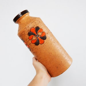 Op de afbeelding: Een cilindervormige, bruine keramische vaas met een getextureerd oppervlak. De vaas heeft een donkerbruine rand en is versierd met oranje en zwarte bloemmotieven. Een decoratief stuk voor in huis.