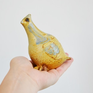 Può includere: Una piccola statuetta di uccello in ceramica, tenuta in mano, caratterizzata da una smaltatura gialla maculata con accenti azzurri sulle ali e sul collo. L'uccello ha un becco appuntito, una piccola cresta e zampe marroni visibili.