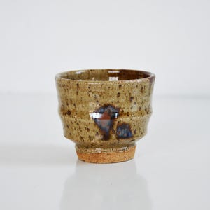 May include: A small, cylindrical ceramic cup with a speckled tan and brown glaze. The exterior features a textured, ridged surface with a prominent dark blue and brown splash pattern. The base is unglazed, revealing a raw, reddish-brown clay texture.