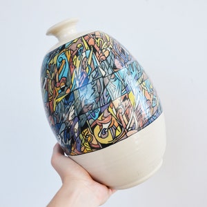 May include: A white ceramic vase with a colourful abstract design. The design features geometric shapes and lines in shades of blue, yellow, orange, and pink.