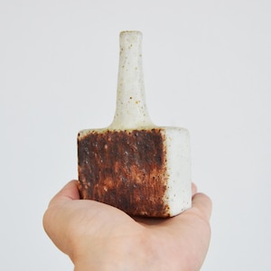 Puede incluir: Un jarrón de cerámica hecho a mano con una base rectangular y un cuello cilíndrico. El jarrón presenta un acabado texturizado blanquecino en el cuello y los lados, con un tono marrón terroso contrastante en la base. El jarrón se sostiene en una mano.