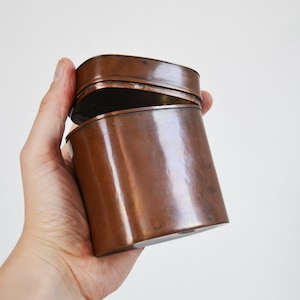 May include: A copper-colored, rectangular tin with a hinged lid, held open. The tin has a slightly rounded shape and a polished finish, reflecting the light. The container is empty.