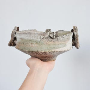 May include: A handcrafted ceramic bowl with a distinctive, textured finish. The bowl has a wide, uneven rim and a flared base. The colour scheme includes shades of brown, grey, and green, with rough, protruding handles.