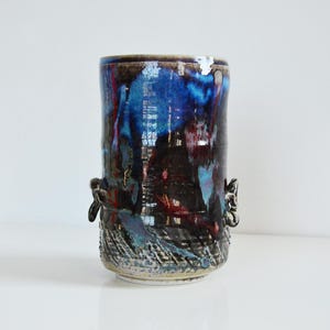 Puede incluir: Un jarrón de cerámica con un esmalte azul oscuro, rojo y negro. El jarrón tiene una forma cilíndrica con una superficie texturizada y dos pequeñas asas a los lados.