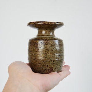 May include: A small, cylindrical ceramic vessel with a speckled, earthy brown and olive green glaze. The object features a distinct, flared rim at the top, a narrow neck, and a wider, straight-sided body with a textured, mottled surface finish.
