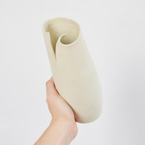 May include: A cream-coloured ceramic vase with a unique, curved design. The vase has a tall, slender shape with a rolled top and a slit down the side. The vase is held in a hand against a white background.
