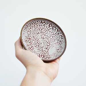 May include: A small, white ceramic bowl with a brown rim and a speckled white glaze. The bowl is held in a hand.