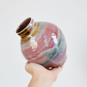 Op de afbeelding: Een ronde keramische vaas met een glanzende afwerking met een mix van roze, blauwe en groene strepen op een beige basis. De vaas heeft een smalle, licht schuine hals met een donkerbruine rand en een glad, bolvormig lichaam dat naar onderen toe loopt.