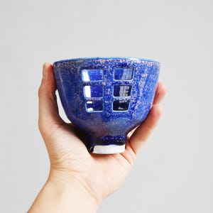 May include: A handmade blue ceramic bowl with a unique square cut-out design. The bowl has a textured surface and a white base. The bowl is held in a hand against a plain background.