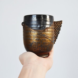 May include: A handmade ceramic cup with a black and gold glaze. The cup has a unique handle made of textured metal.