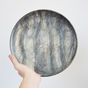 May include: A round ceramic plate with a blue and white glaze. The plate has a design of three figures carved into the surface.
