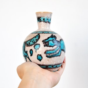 May include: A small, round ceramic vase with a beige speckled finish. The vase features turquoise and black abstract designs. The rim and neck are a lighter beige colour. The vase is held in a hand.