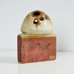 May include: A handcrafted ceramic sculpture with a rectangular base and a rounded top. The base is a reddish-brown colour with a textured surface, while the top is off-white with three circular openings. The sculpture has a rustic, earthy aesthetic.