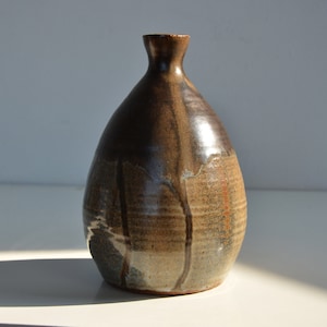 Puede incluir: Un jarrón de cerámica marrón con cuello estrecho y cuerpo redondeado. El jarrón tiene un esmalte moteado con tonos marrones, grises y blancos.