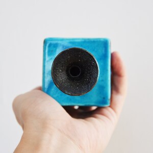 K&ouml;nnte beinhalten: Ein blauer Kerzenhalter aus Keramik mit einem schwarzen Metalldeckel. Der Halter ist quadratisch und hat eine glatte, gl&auml;nzende Oberfl&auml;che.