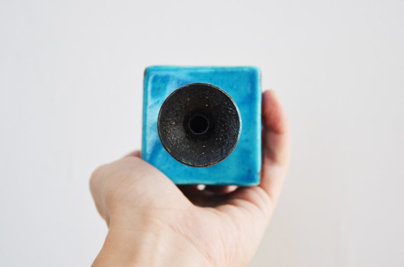 K&ouml;nnte beinhalten: Ein blauer Kerzenhalter aus Keramik mit einem schwarzen Metalldeckel. Der Halter ist quadratisch und hat eine glatte, gl&auml;nzende Oberfl&auml;che.