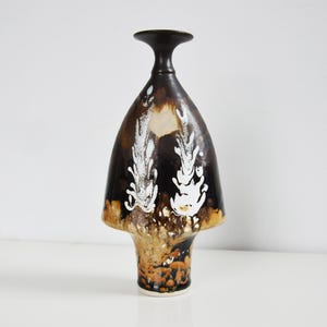 Op de afbeelding: Een hoge, decoratieve keramische vaas met een donkerbruine, uitlopende body en een smalle hals. De vaas heeft witte, abstracte bloemmotieven op de voorkant. De basis is lichtbruin met oranje accenten. De vaas is ongeveer 30 cm hoog.