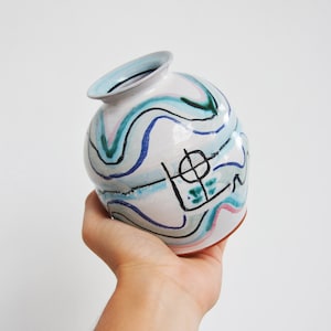 Puede incluir: Un jarrón de cerámica blanca con diseños abstractos azules, verdes y rosas. El jarrón tiene un cuerpo redondo y una abertura ligeramente acampanada.