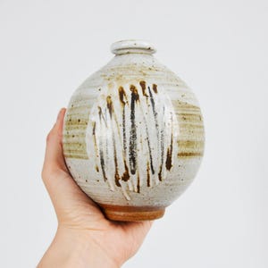 Op de afbeelding: Een witte keramische vaas met bruine en zwarte glazuuraccenten. De vaas heeft een ronde vorm en een kleine opening aan de bovenkant.