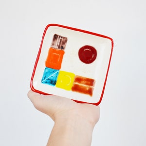 May include: A small, square ceramic dish with a red rim. The dish is decorated with a geometric pattern of colorful squares and a red circle.