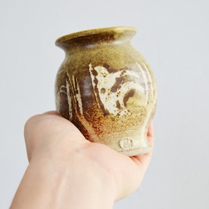 May include: A small, brown and white ceramic vase with a unique design. The vase has a rounded shape and a slightly flared opening.